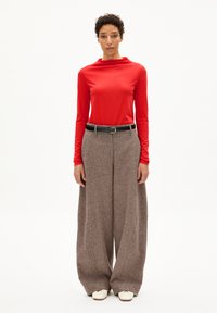 Roterlangärmliger Oberteil mit hohem Kragen, kombiniert mit weiten braunen Textilhosen und einem schwarzen Gürtel. Weiße Schuhe vollenden den Look.