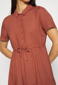 Robe courte de couleur rouille à manches courtes, avec un devant à boutons, un col, et une ceinture à nouer pour un ajustement réglable. Tissu lisse et fluide.