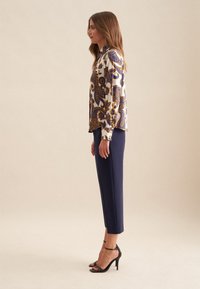 Blusa a maniche lunghe color crema con motivi floreali blu e oro, abbinata a pantaloni tailored blu navy e tacchi alti neri con cinturini.