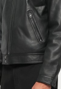 Juoda odinė striukė su užtrauktuku užsegama kišene, sklandžiu paviršiumi, užsegimu mygtukais apatine dalimi ir prigludusia forma.