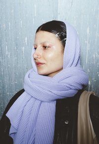 Sciarpa lavorata a maglia color lavanda avvolta intorno al collo, con una finitura testurizzata. Giacca di pelle nera visibile sulla spalla, con un accessorio marrone chiaro.