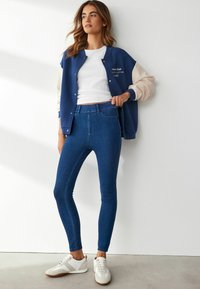Námořnická a béžová varsity bunda se cvoky, bílá tričko, modré skinny džíny a bílé tenisky s hnědou podrážkou.