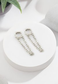 Silverfärgade hoopsringar med länkade kedjedroppar. Slät metallfinish, rund form, modern design, visad på en vit rund pedistal.