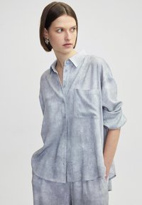 Camisa de hombre azul claro, texturizada, con cuello, bolsillo en el pecho y mangas enrolladas. Tela suave con un patrón sutil. Corte casual.