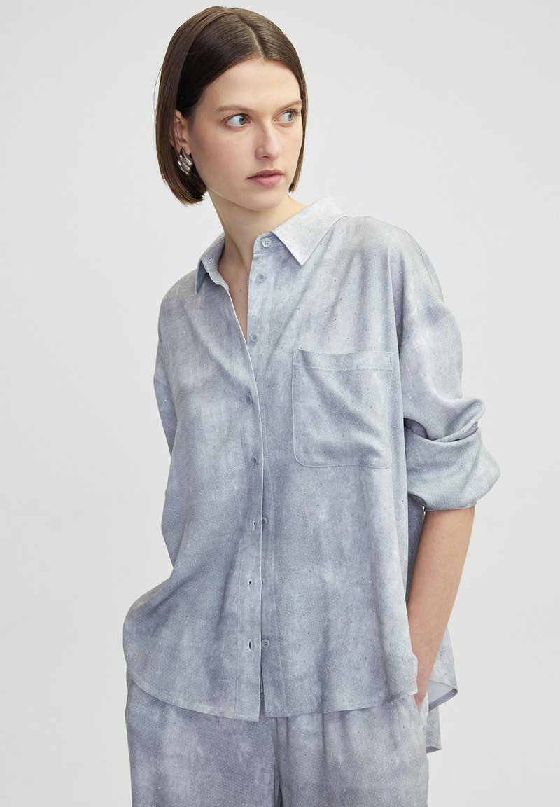Camisa de hombre azul claro, texturizada, con cuello, bolsillo en el pecho y mangas enrolladas. Tela suave con un patrón sutil. Corte casual.