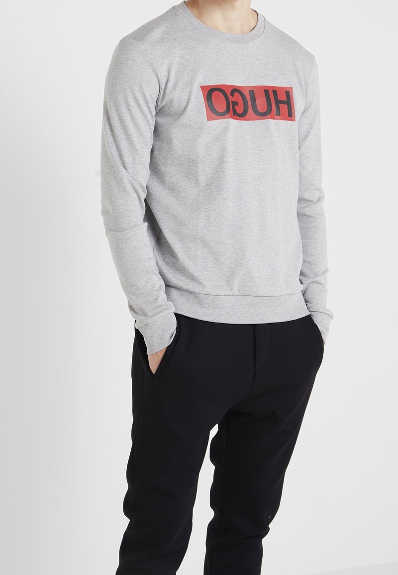 Homme portant un sweat-shirt gris clair avec un logo rectangulaire rouge et un pantalon noir, mains dans les poches, sur fond blanc.