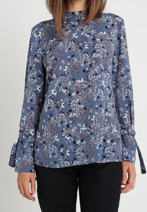 Niebieska bluzka z długim rękawem z białym, czarnym i rdzawym kwiatowym wzorem, z wiązaniami przy mankietach, noszona z czarnymi spodniami.