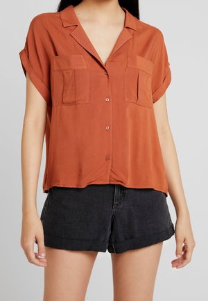 Mujer que lleva una camisa suelta de manga corta color óxido con dos bolsillos en el pecho y pantalones cortos de mezclilla negros con dobladillo enrollado.