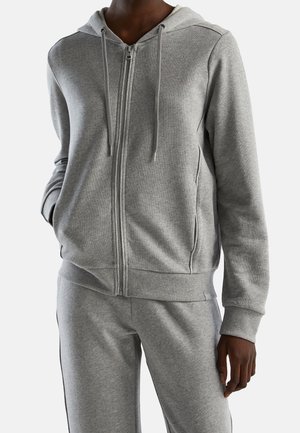 Person iført en grå lynlåshoodie med snore og matchende grå joggingbukser mod en ensfarvet hvid baggrund.
