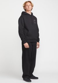 Sweat à capuche noir avec une poche avant et un cordon de serrage. Associé à un pantalon large noir et des baskets sombres. Détail de logo minimaliste sur la poitrine.