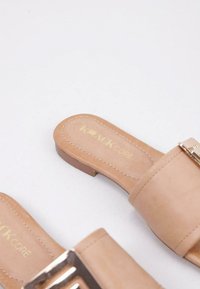 Chanclas deslizantes beige hechas de material sintético, con suela plana, tiras anchas y detalles de hebilla metálica. Logotipo estampado en la plantilla.
