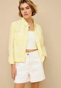 Žltá košeľa na gombíky s dvoma náprsníkovými vreckami, oblečená cez biele crop top. Skombinovaná s bielymi šortkami, čo ukazuje uvoľnený outfit.