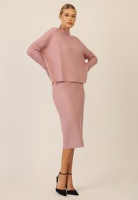 Rosa Strickpullover mit gerippten Ärmeln, kombiniert mit einem passenden, taillierten Bleistiftrock, beide mit glatter Textur und minimalistischem Design. Schwarze High Heels.