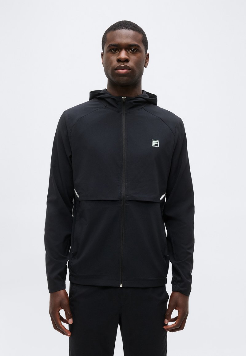 Νέος άνδρας που φοράει μαύρο φλις με κουκούλα και φερμουάρ και μικρό λευκό λογότυπο στο στήθος, στέκεται μπροστά σε λευκό απλό φόντο.