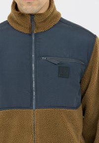 Fleecejacka med en marinblå ovandel och en dragkedjeförsedd bröstficka, samt en nedre del i brun, fluffig fleece. Ståkrage och diskret varumärkesdetalj.