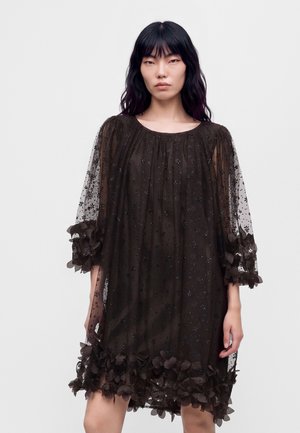 Un mannequin se tient devant un fond clair, vêtu d’une robe noire ample et transparente, ornée de détails scintillants et dotée de poignets et d’un ourlet volantés.