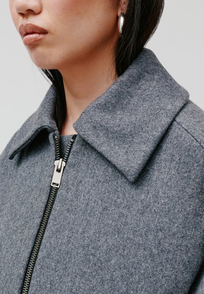 Grau melierte Wolljacke mit einem Kragen, versehen mit einem silbernen Reißverschluss und einer strukturierten Oberfläche. Akzente bilden große Creolen.