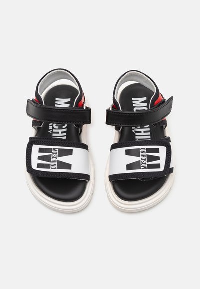 Sandalias en azul marino y blanco con correas ajustables, que cuentan con un gran logo negro de Moschino, suela negra y acentos rojos. Diseño plano, material sintético.