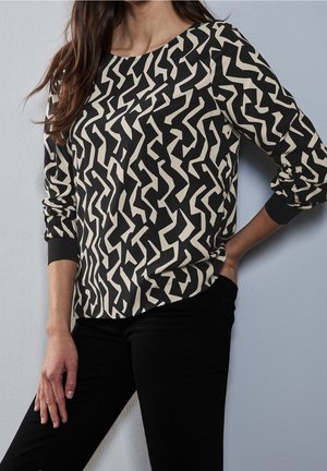 Frau trägt ein langärmeliges schwarz-beige geometrisch gemustertes Oberteil mit schwarzen Hosen, steht vor einer hellgrauen Wand.