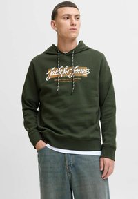 Mørkegrøn hættetrøje med hvid og orange grafisk tryk, justerbar snor og ribbede mansjetter. Bæres over en hvid t-shirt.