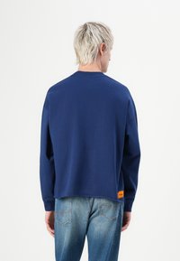 Mørkeblå genser med lange ermer, rund hals, litt cropped design og en oransje etikett på venstre side. Strukturert stoff.