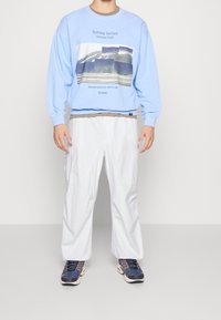 Ljusblå sweatshirt med grafisk design, rundhals, och långa ärmar, i kombination med vita cargobyxor och marinblå sneakers med accenter.