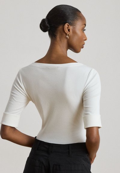 Femme avec les cheveux en chignon porte un haut blanc à manches trois-quarts et un pantalon noir, de profil sur un fond uni.
