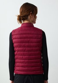 Gilet imbottito bordeaux con trapuntatura orizzontale, tessuto leggero, collo rotondo e design aderente, indossato sopra una maglietta nera a maniche lunghe.