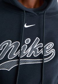 Svart hoodie med en stor grå "Nike"-logotyp som har vita accenter, texturerade detaljer och dragsnören vid halsringningen.