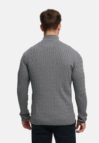 Grå kabelstrikket genser med høy krage, som har teksturerte mønstre over hele og ribbestrikkede mansjetter og kant. Sett bakfra.