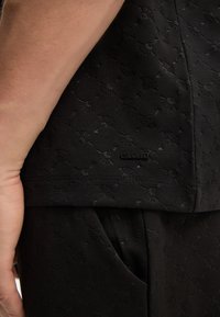Nahaufnahme eines schwarzen, strukturierten Stoffes mit einem erhabenen Muster aus blumigen Designs. Enthält ein kleines schwarzes Etikett mit dem Markennamen am Saum.