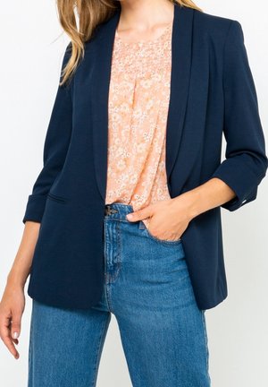 Kvinde iført navy blazer med opkørte ærmer, ferskenfarvet blomsterbluse og blå højtaljede jeans, hånd i lommen, mod hvid baggrund.