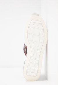 Sneaker bianco con pannelli laterali in tessuto rosso e nero, suola in gomma con un motivo a griglia e finitura liscia. La suola è di un grigio chiaro.
