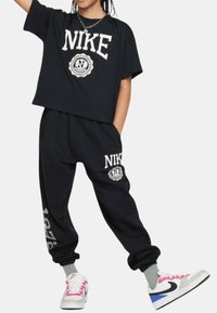 Camiseta negra de Nike corta y pantalones de chándal a juego, con estampados de logotipos en blanco. Las zapatillas tienen acentos rosas con detalles en gris y negro.