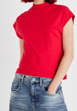 Henkilö, joka pukeutunut punaiseen lyhythihaiseen yläosaan, joka on puettu valkoista taustaa vasten vaaleansinisiin farkkuihin ja jolla on toinen käsi taskussa.