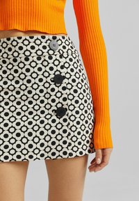 Svart och vitt mönstrad minikjol med cirkulära mönster, med svarta knappar på sidan och ett slätt tyg. Tillsammans med en ribbad orange topp.