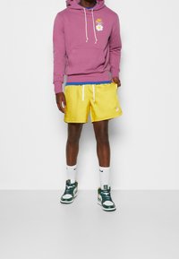 Fialová mikina s kapucí a přední kapsou, zdobená grafikou sedmikrásky, v kombinaci se žlutými šortkami s modrým pasem. Oblečeno s bílými ponožkami a zelenými teniskami.