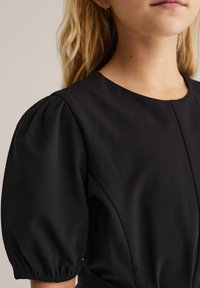 Crna haljina s okruglim izrezom, kratkim napuhnuti rukavima i glatkom teksturom tkanine. Sadrži uski dio tijela s suptilnim nabranim detaljima.