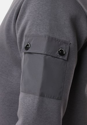 Gray long-sleeve shirt features a contrasting gray fabric patch on the sleeve with two black snap buttons and a logo detail.
