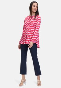 Maglietta a maniche lunghe svasata con motivo geometrico in rosso e rosa. Tessuto liscio, vestibilità comoda. Abbinata a pantaloni slim blu navy e sandali bianchi con tacco a blocco.
