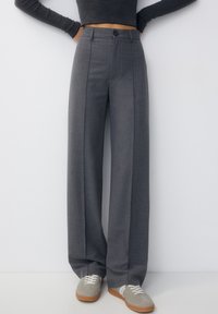 Persoon in hoog-gesneden, brede grijze broek met beige sneakers en een korte zwarte lange mouw top, met handen op de heupen.