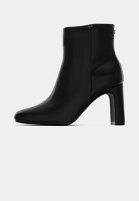 Černá kožená kotníková bota s špičatou špičkou a blokovým podpatkem, hladký povrch, boční zip uzavřený, izolováno na bílém pozadí.