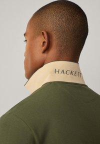 Maslinasto zelena polo majica s svijetlosmešavim ovratnikom na kojem je izvezeno "HACKETT", s kratkim rukavima i teksturiranim materijalom.