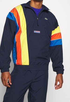 Mann trägt eine marineblaue Lacoste-Trainingsanzugjacke mit gelben, orangenen und blauen diagonalen Streifen und einem Kragen mit Viertelreißverschluss.