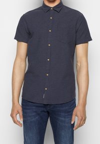 Camicetta blu navy a maniche corte con un motivo testurizzato, chiusura a bottoni e una tasca singola sul petto; realizzata in tessuto leggero.