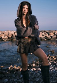 Sweat-shirt à capuche gris et jupe-culotte imprimée, tous deux avec une coupe ample. Modèle portant des bottes en caoutchouc noires jusqu'au genou. Côte rocheuse naturelle en arrière-plan.