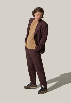 Traje burdeos con chaqueta a medida y pantalones a juego, combinado con una camiseta beige y zapatillas con patrón. Tejido suave, corte relajado.