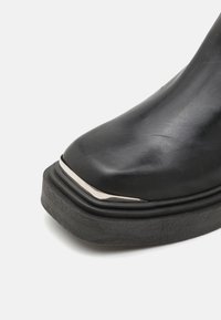 Bota de cuero negra hasta el tobillo con puntera redondeada, que presenta un acento metálico cerca de la punta y una suela negra gruesa y texturizada.