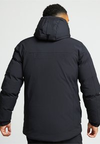 Melna pūkaina jaka ar kapuci, ar gludu tekstūru, elastīgiem piedurknēm un aizmugures ventilācijas detaļu. Materiāls izskatās ūdensizturīgs.