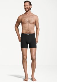 Boxer brevi neri con un design aderente e una fascia elastica in vita, indossati da un uomo in piedi a piedi nudi su uno sfondo neutro.